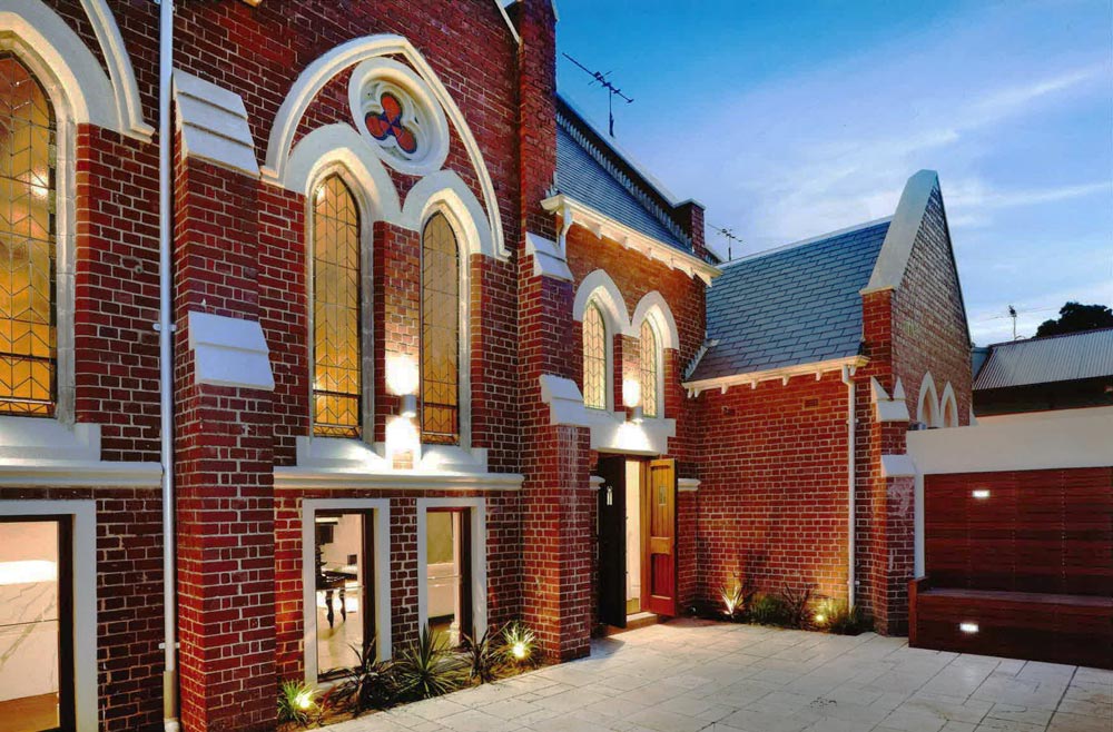 Red brick building with tall arched stained glass windows, white trim, and pointed rooflines. Outdoor lights illuminate the entrance and surrounding courtyard at dusk.